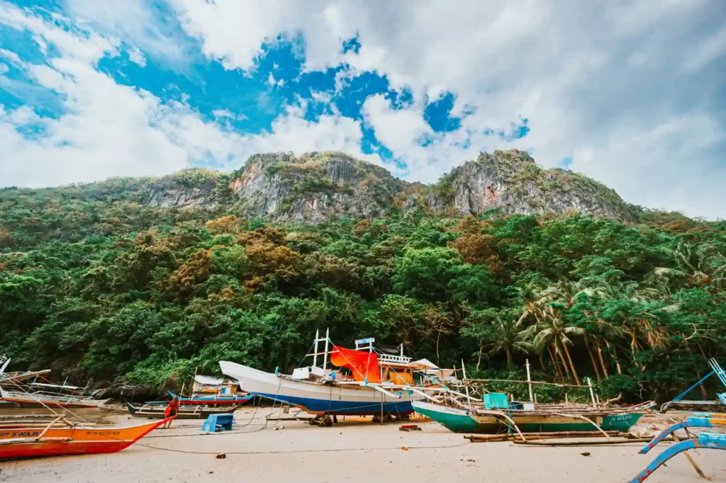 Traditional outrigger boats resting on a sandy beach below forested limestone cliffs in Bohol, Philippines.