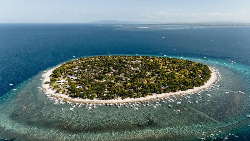 Aerial view of a small tropical island near Bohol surrounded by coral reefs, clear blue water, white beaches, and traditional boats.