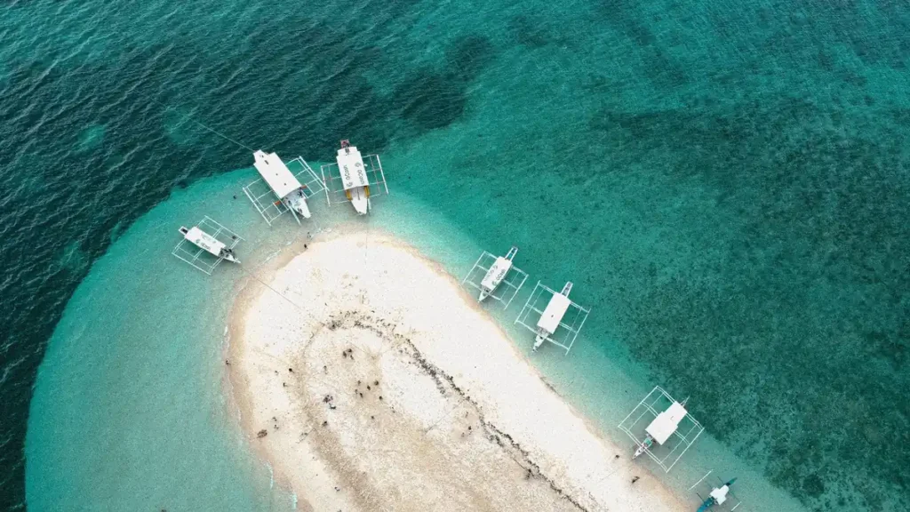 Aerial view of a white sandbar in Bohol surrounded by clear turquoise water, with traditional outrigger boats anchored around the shore.