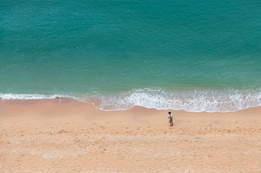 Aerial view of turquoise sea waves meeting a quiet sandy beach with one person walking along the shoreline.