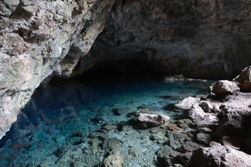 Crystal-clear blue water inside a natural limestone cave, surrounded by rugged rock formations and submerged stones, creating a tranquil underground lagoon scene.