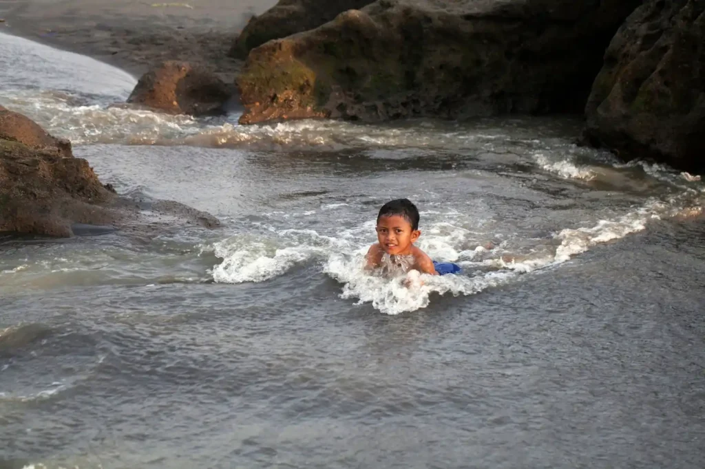 Child playing in a natural tidal pool between coastal rocks as seawater flows through a beach cove during low tide.
