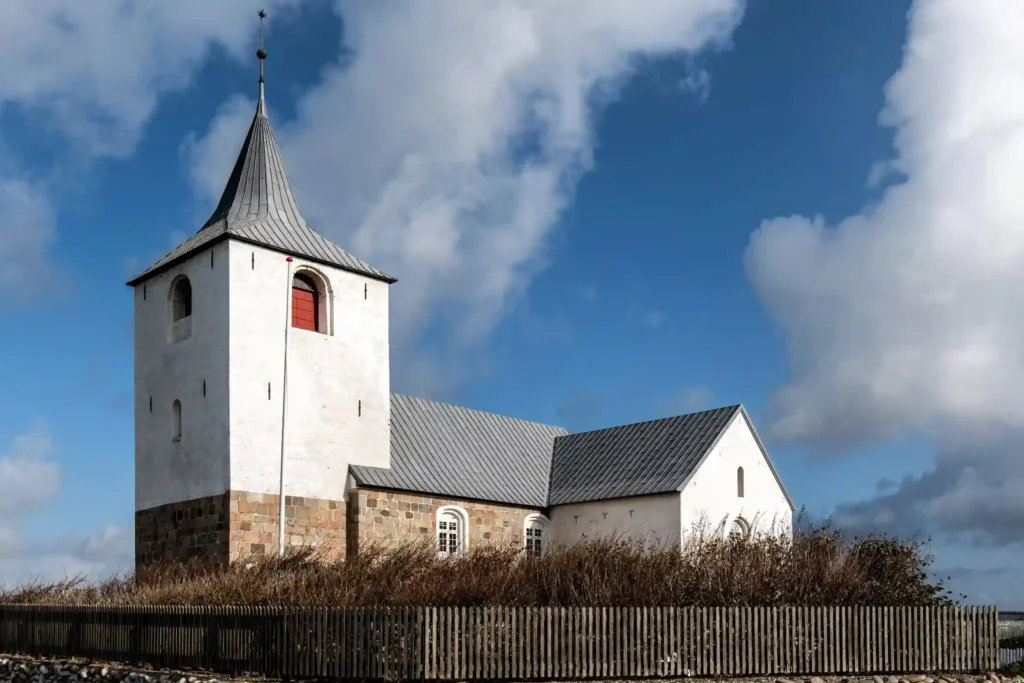 Traditional white stone church with a tall steeple and gray roof, surrounded by a wooden fence under a dramatic blue sky with large clouds.