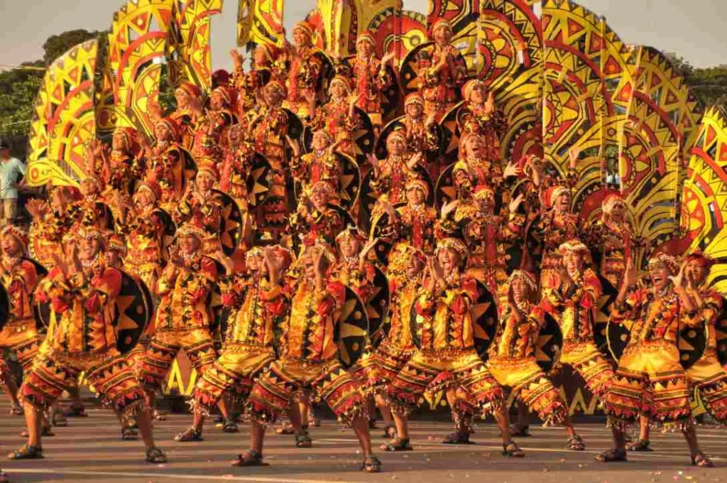A large group of cultural festival dancers in ornate yellow and red patterned costumes performing outdoors.