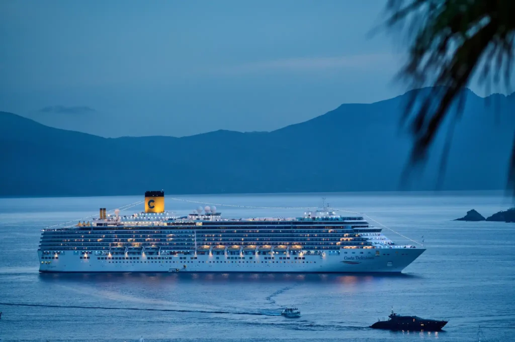 Illuminated cruise ship sailing across calm blue waters at dusk with mountain silhouettes in the background.