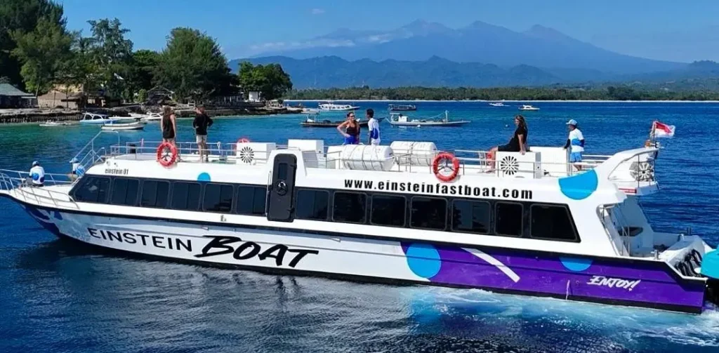 Fast boat traveling near the Gili Islands with passengers on board, blue sea, coastal village, and mountains in the background.