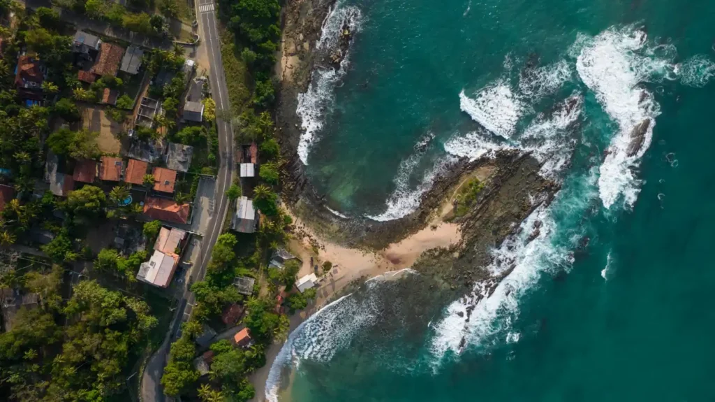 Aerial view of Goyambokka Beach in Sri Lanka with turquoise water, rocky coastline, waves, palm trees, and a coastal road.