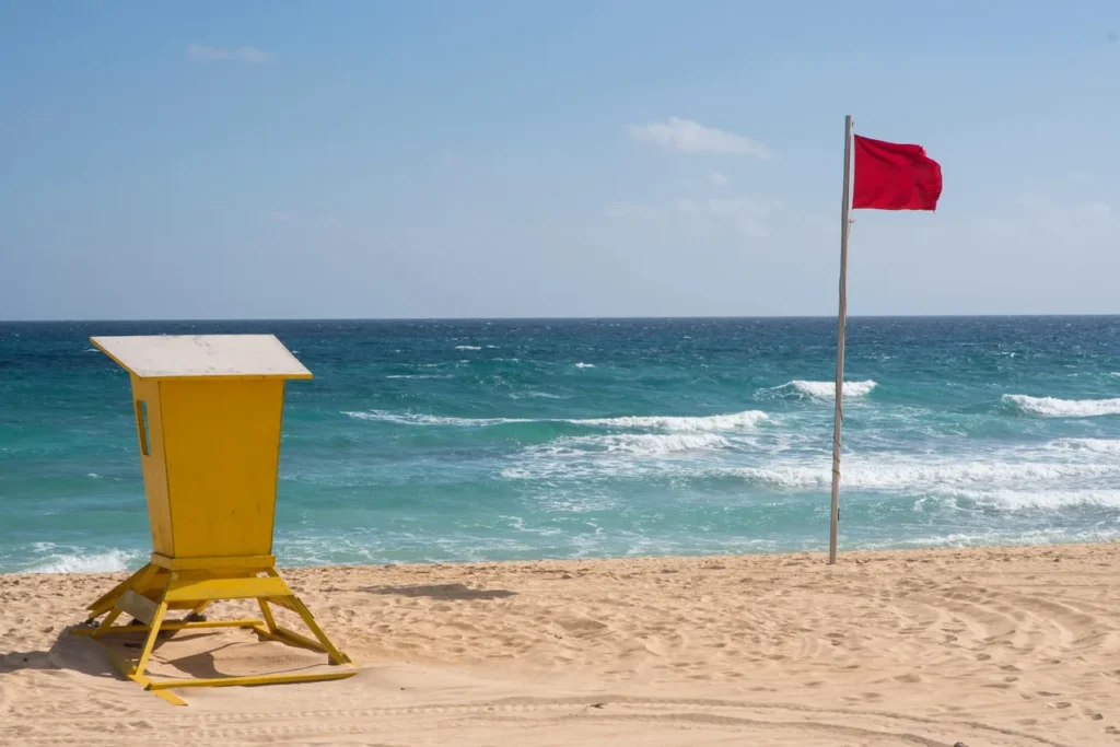 Red warning flag and lifeguard hut on a sandy beach with rough sea waves, showing unsafe swimming conditions near Goyambokka Beach.