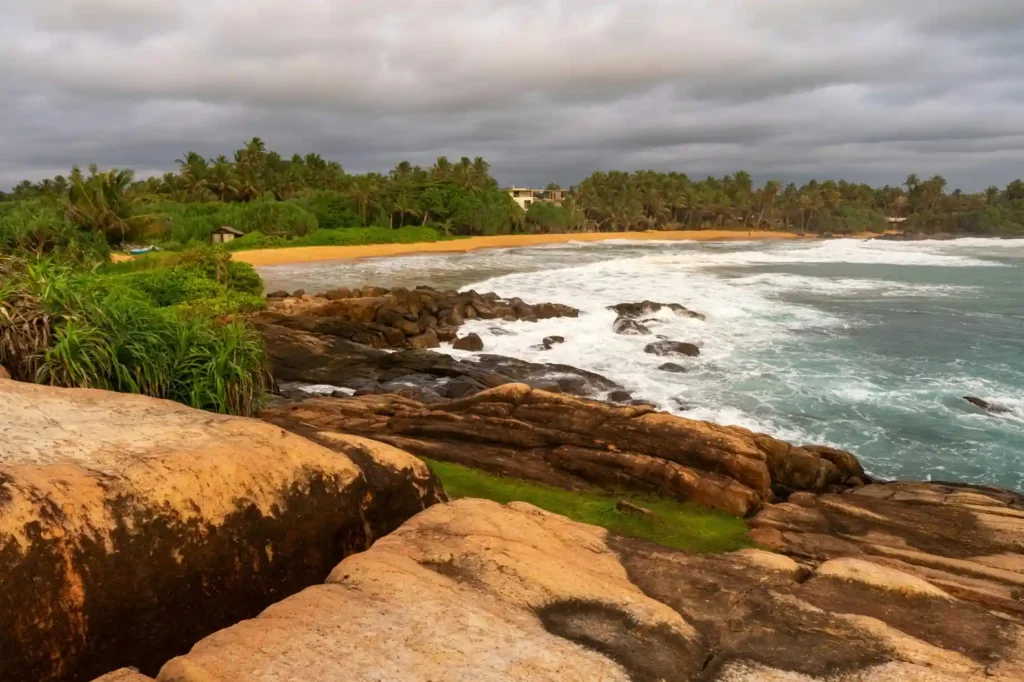 Rocky coastline at Goyambokka Beach in Sri Lanka with golden sand, palm trees, cloudy sky, and waves hitting the shore.
