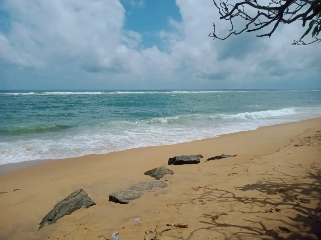 Quiet tropical beach shoreline with turquoise waves, golden sand, rocks, and tree shadows under a cloudy blue sky.