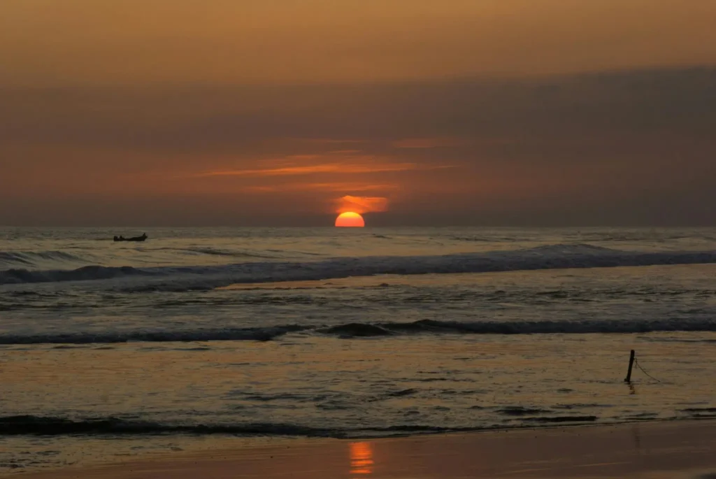 Sunset over the ocean near Goyambokka Beach with orange sky, gentle waves, and a small boat silhouette on the horizon