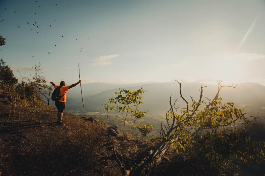 Hiker standing on a mountain ridge at sunrise overlooking a scenic valley and distant mountain ranges.