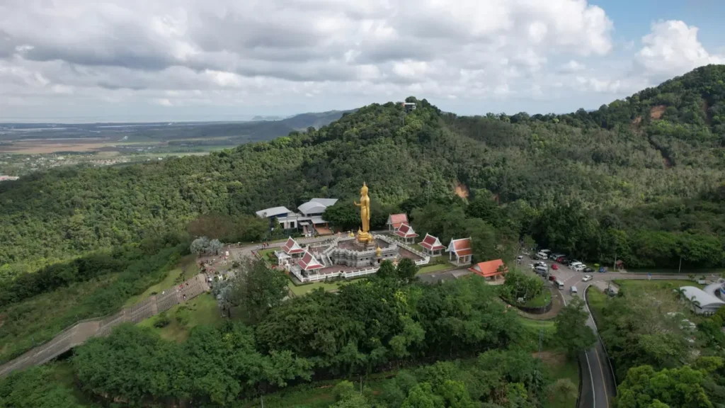 Aerial view of a golden Buddha statue and temple complex on a green hilltop with mountains and cloudy sky.