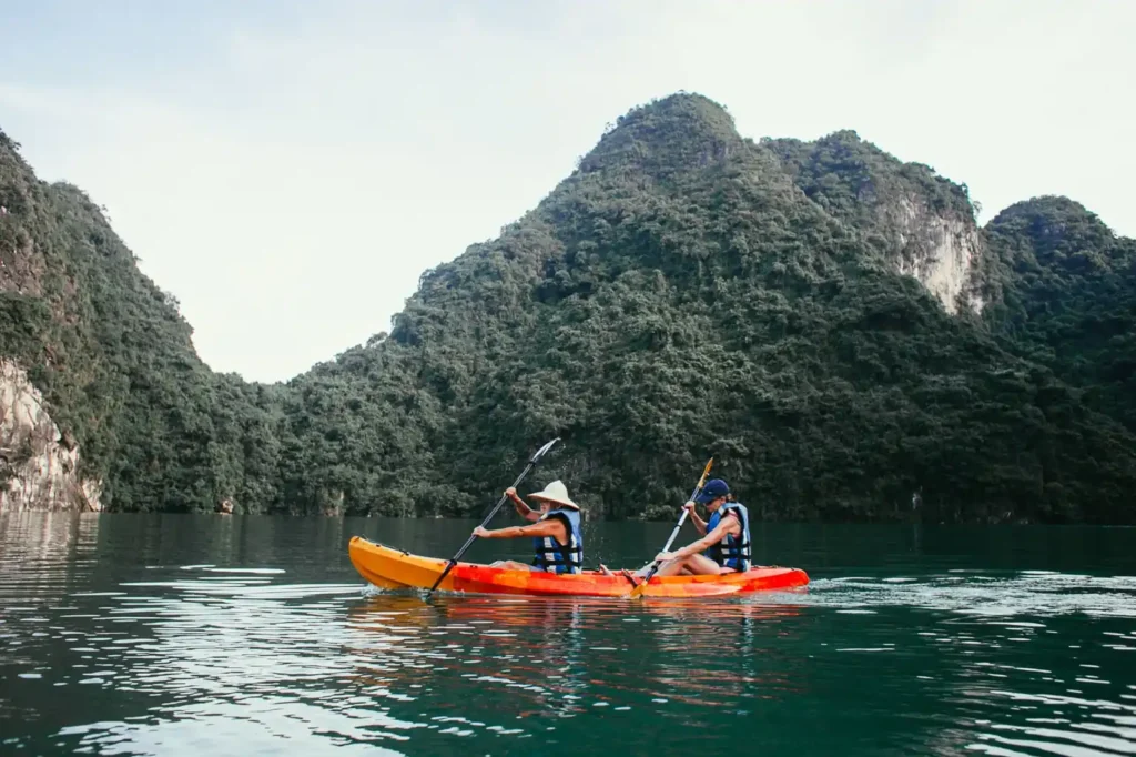 Two people kayaking on a calm lagoon surrounded by lush green limestone cliffs.