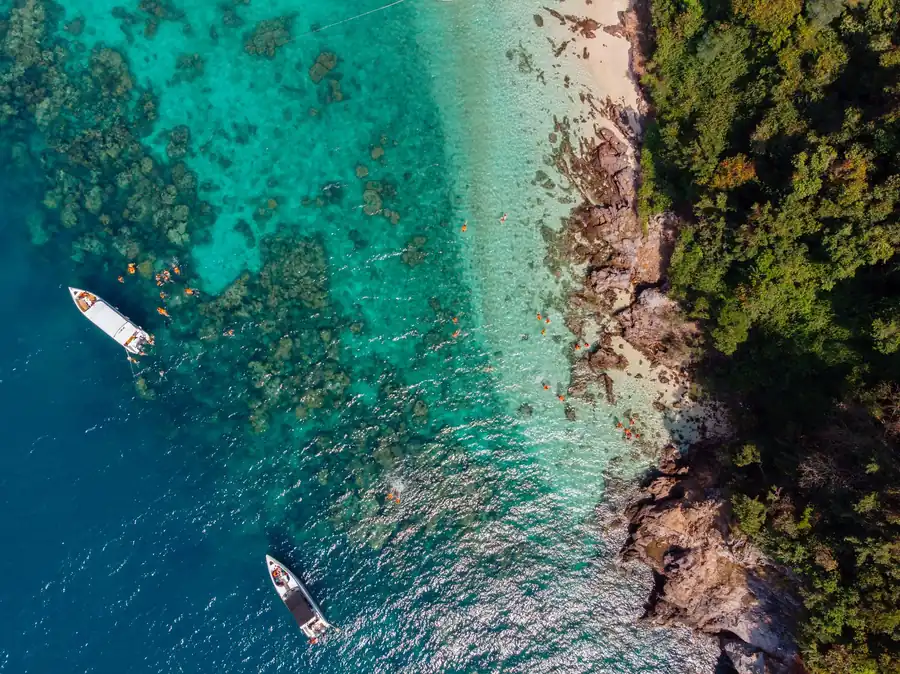 Aerial view of turquoise Koh Lipe water with boats, snorkelers, coral reefs, rocky shoreline, and dense green island forest.