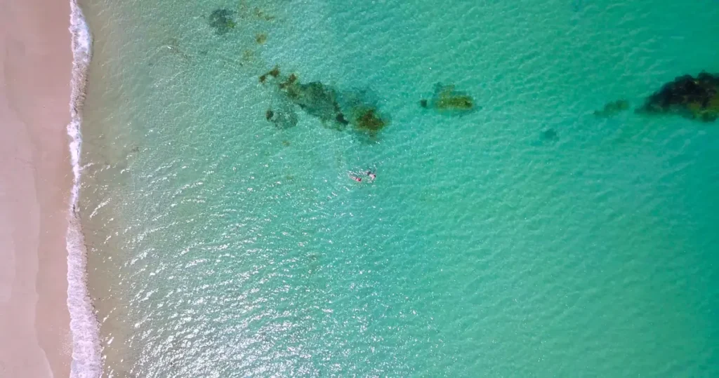 Aerial view of a snorkeler swimming in clear turquoise water beside a sandy Koh Lipe shoreline and shallow reef rocks. 