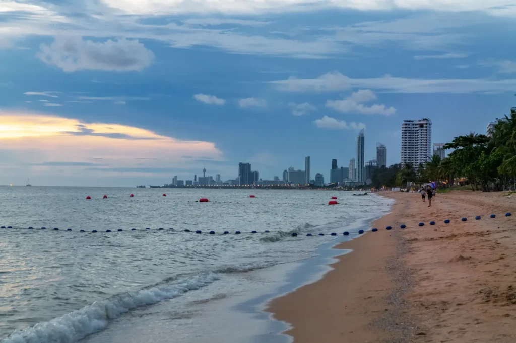 Beach shoreline at sunset with gentle waves, city skyline, palm trees, and evening clouds in the distance.
