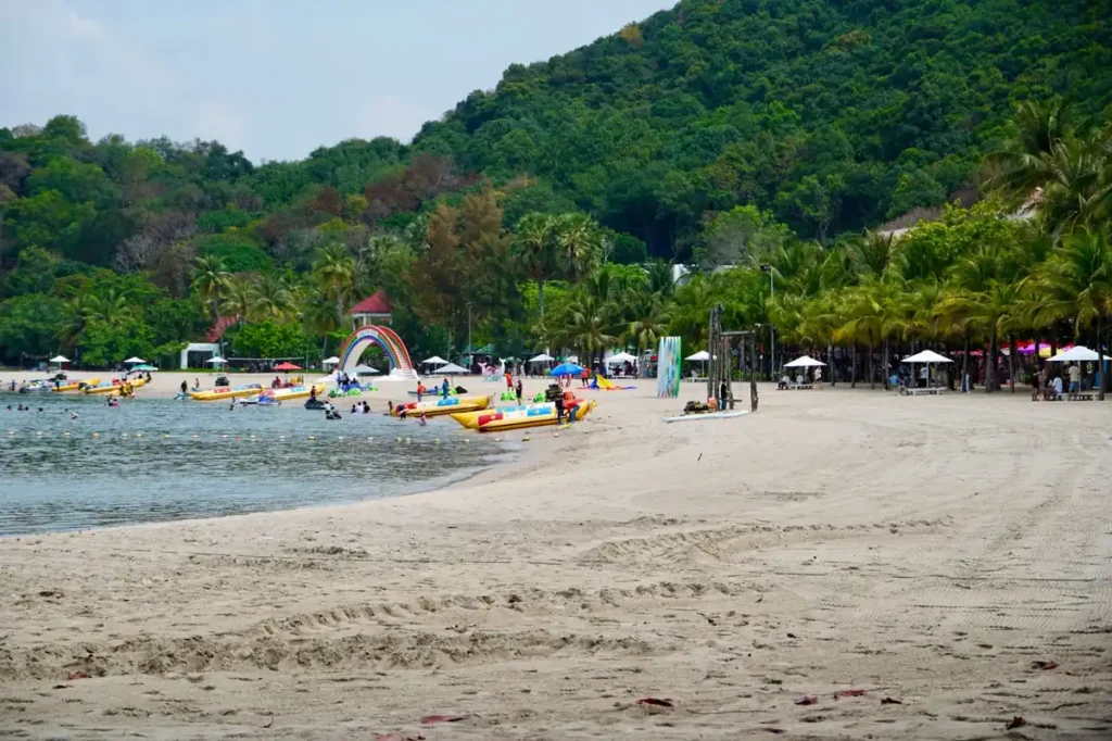 Busy tropical beach with water activities, palm trees, visitors, and a forested hillside in the background.