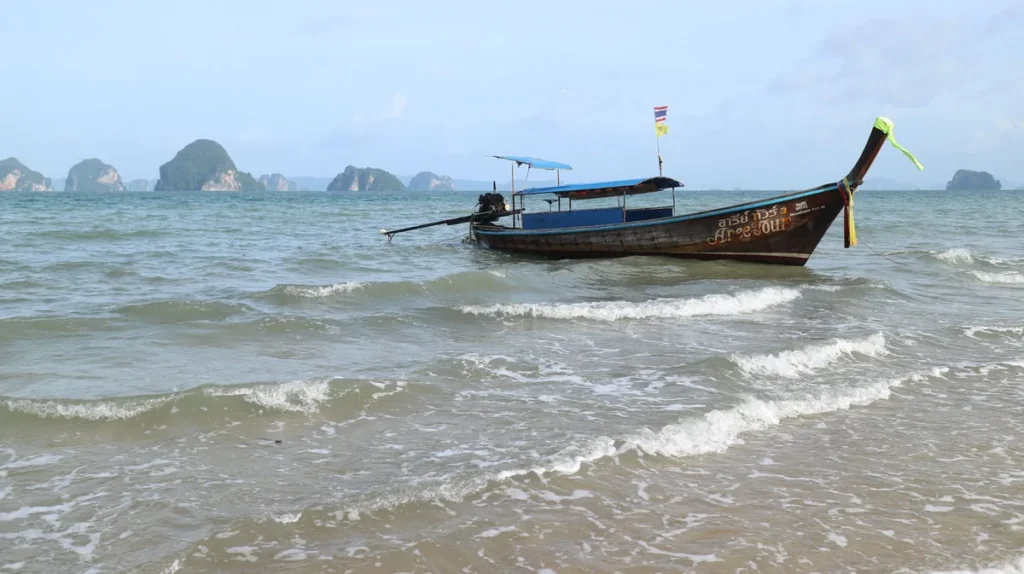 Traditional Thai longtail boat floating near the shore with small waves and limestone islands in the background.