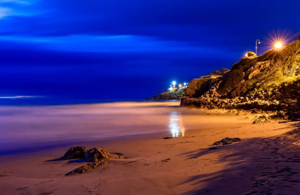 Quiet tropical beach at night with illuminated rocky cliffs, calm water, and lights reflecting across the shoreline.