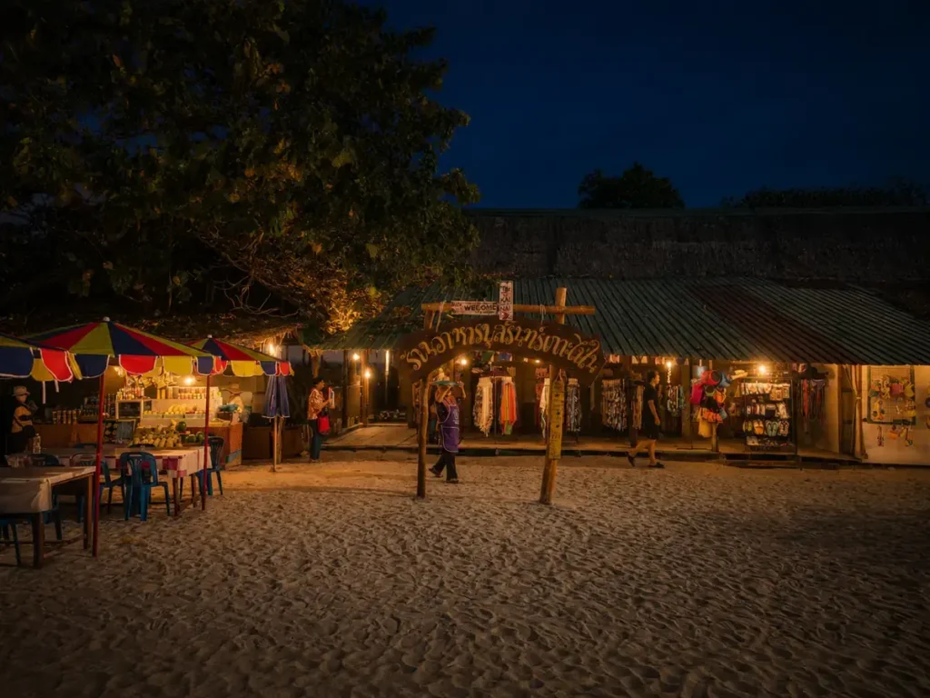 Koh Lipe beach market at night with warm shop lights, colorful umbrellas, souvenir stalls, and visitors on the sand.