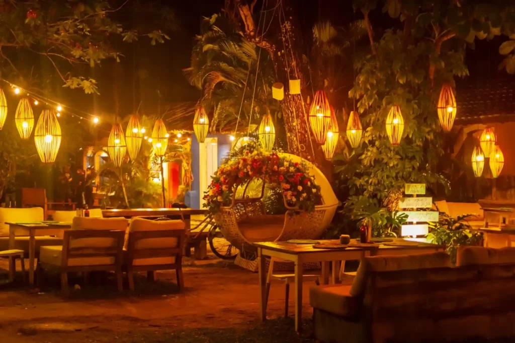 Warmly lit tropical beach bar at night with hanging lanterns, outdoor seating, flowers, and palm trees.