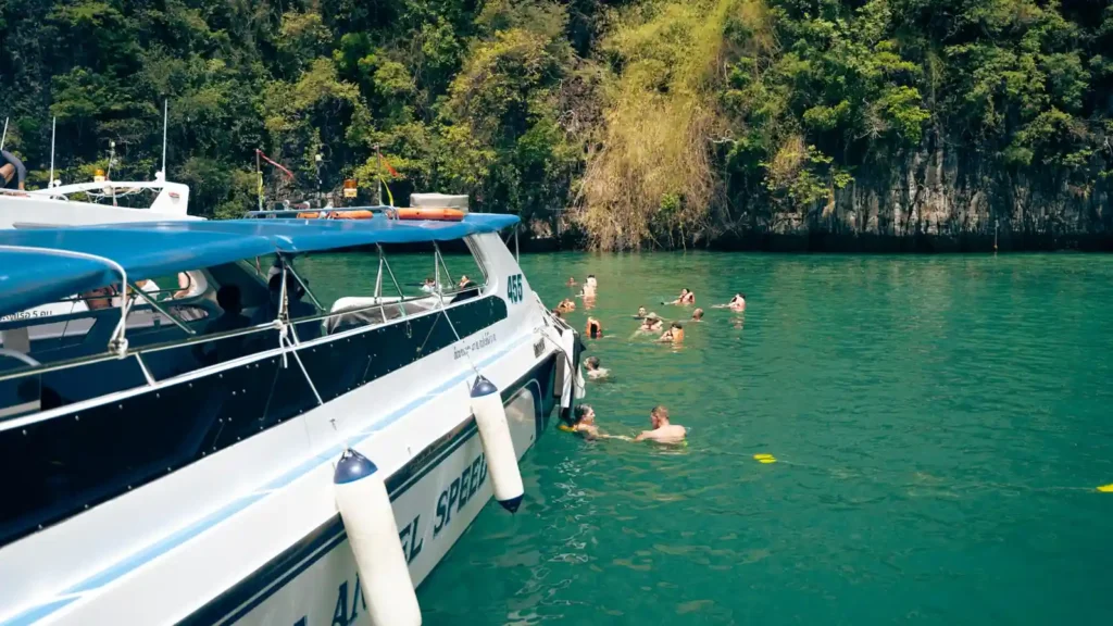 Travelers swimming beside a speedboat in clear green water near the forested coast of Koh Lipe, Thailand. 