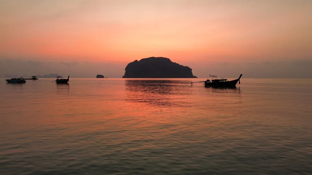 Sunset over calm Koh Lipe waters with longtail boats and a dark island silhouette in the distance.