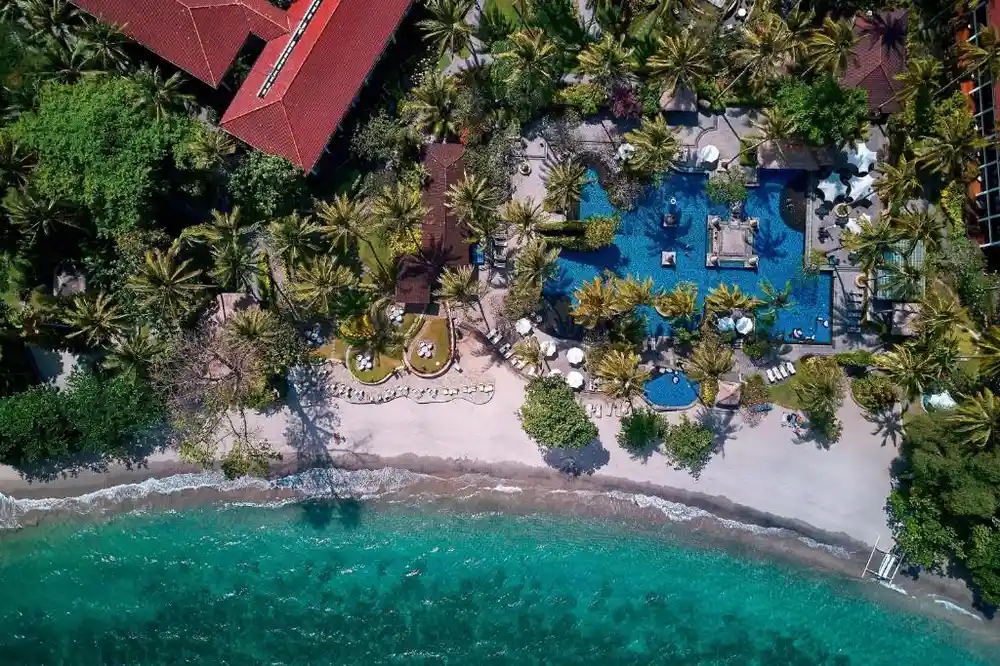 Aerial view of a beachfront resort in Lombok with palm trees, swimming pools, sandy beach, and turquoise water.
