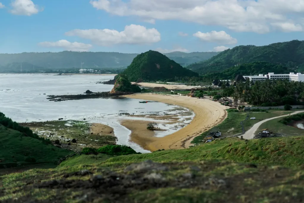 Panoramic view of a quiet Lombok beach with green hills, calm water, sandy shoreline, and coastal resorts under a cloudy blue sky.