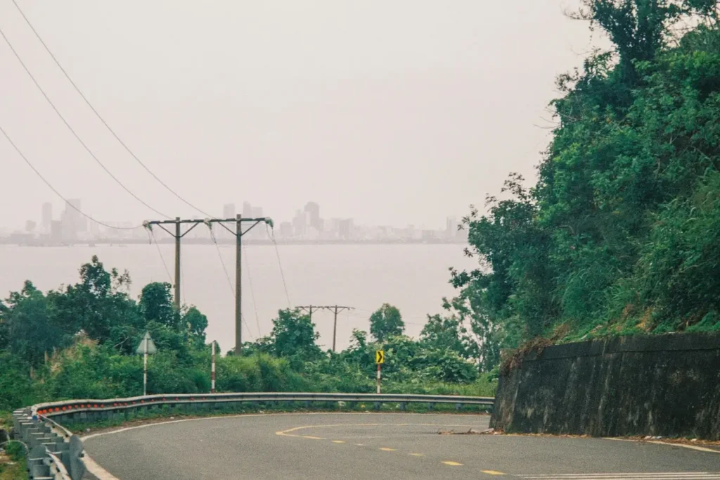 Curved coastal road in Lombok with green hills, power lines, and hazy ocean views leading toward hidden beaches.