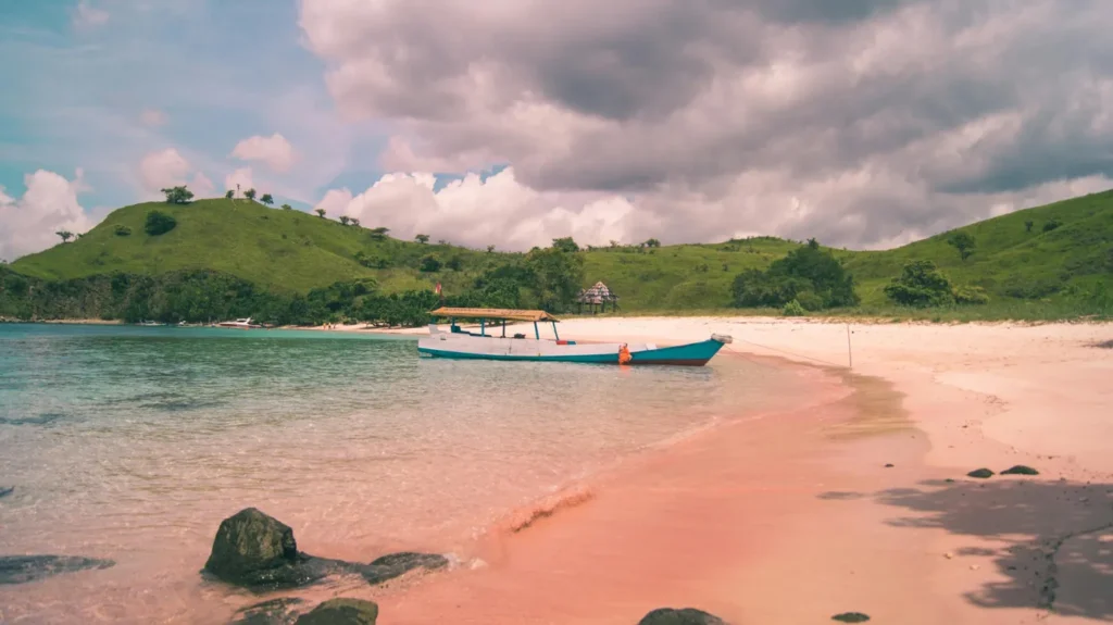 Quiet hidden beach in Lombok with pink sand, clear shallow water, a small boat, green hills, and cloudy sky.