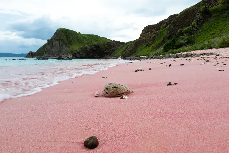 Pink sand beach in Lombok with gentle waves, rocky cliffs, green hills, and scattered stones along the shore.