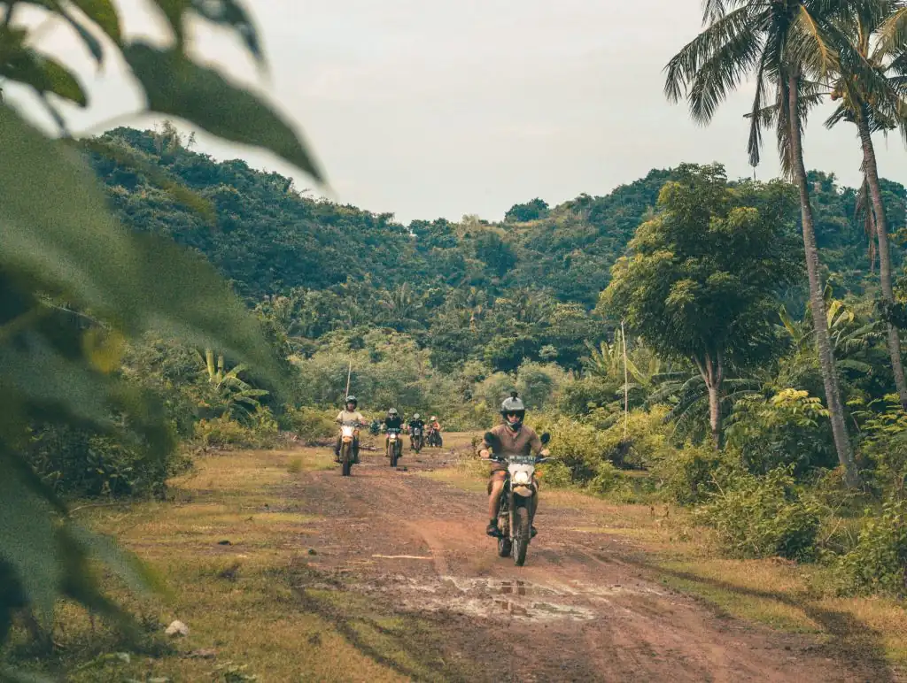 Scooter riders traveling along a muddy dirt road surrounded by tropical greenery and palm trees in Lombok.