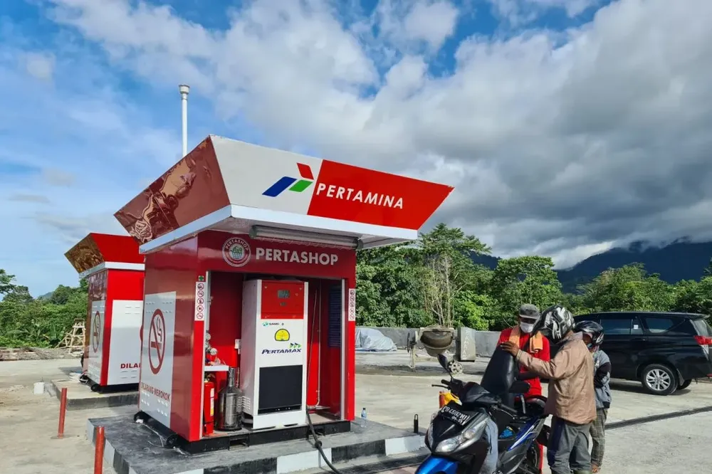 Scooter rider refueling at a Pertamina fuel station in Lombok with green mountains and cloudy sky in the background.