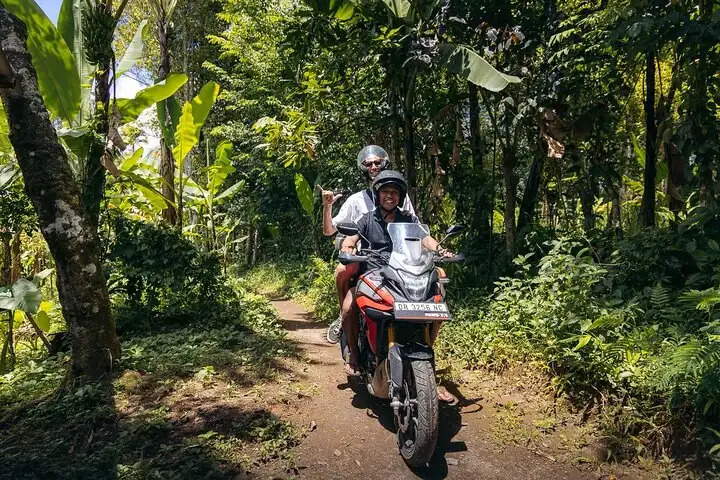 Two travelers riding a scooter through a lush green jungle road in Lombok.