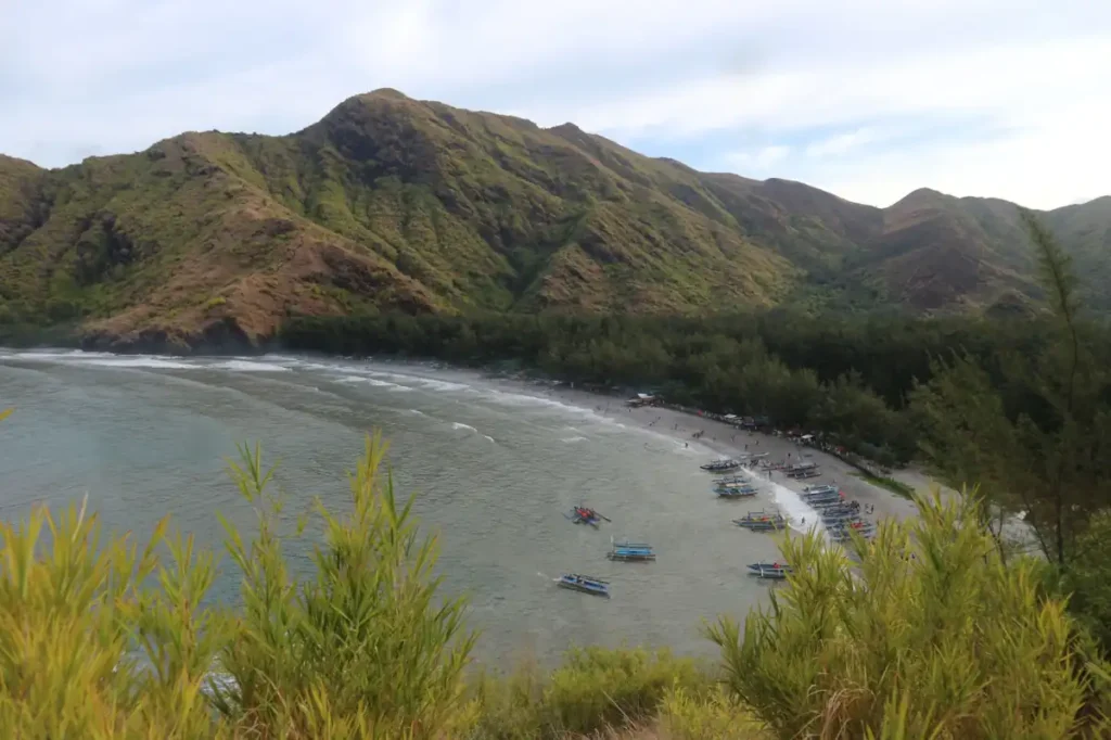 Coastal beach backed by green mountains with traditional boats along the shoreline.