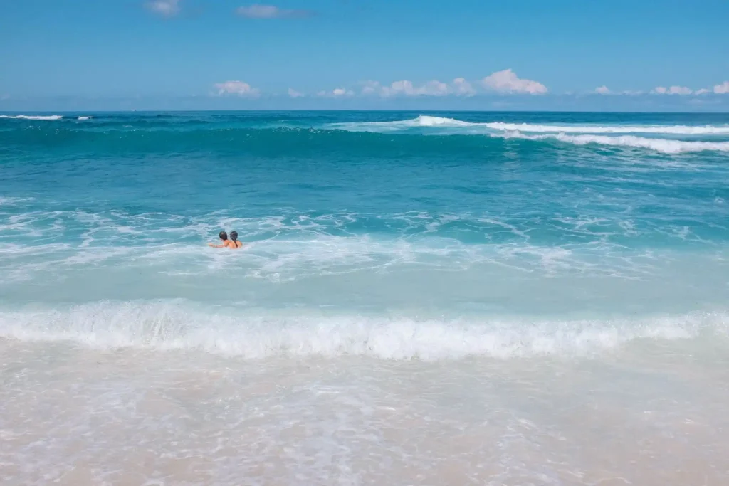Two people swimming in clear turquoise ocean water near a sandy beach, with rolling waves, white surf, and a bright blue sky in a tropical coastal setting.