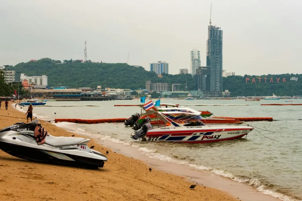 Pattaya Beach shoreline with speedboats, jet ski, city skyline, and hillside Pattaya sign in the background. 