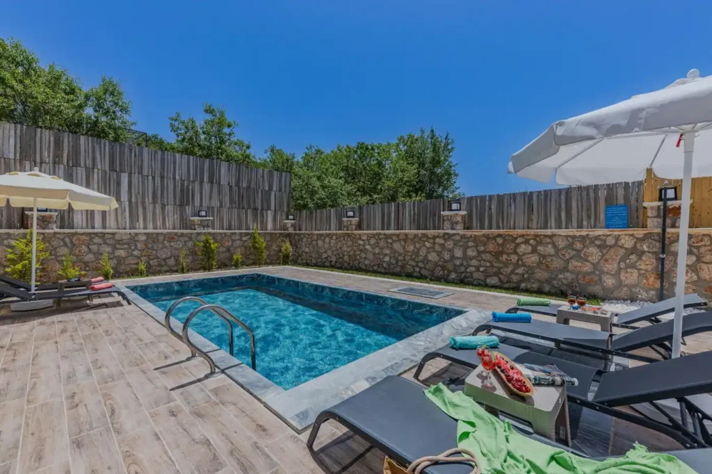Private villa pool with sun loungers, white umbrellas, stone walls, blue sky, and towels beside the water.