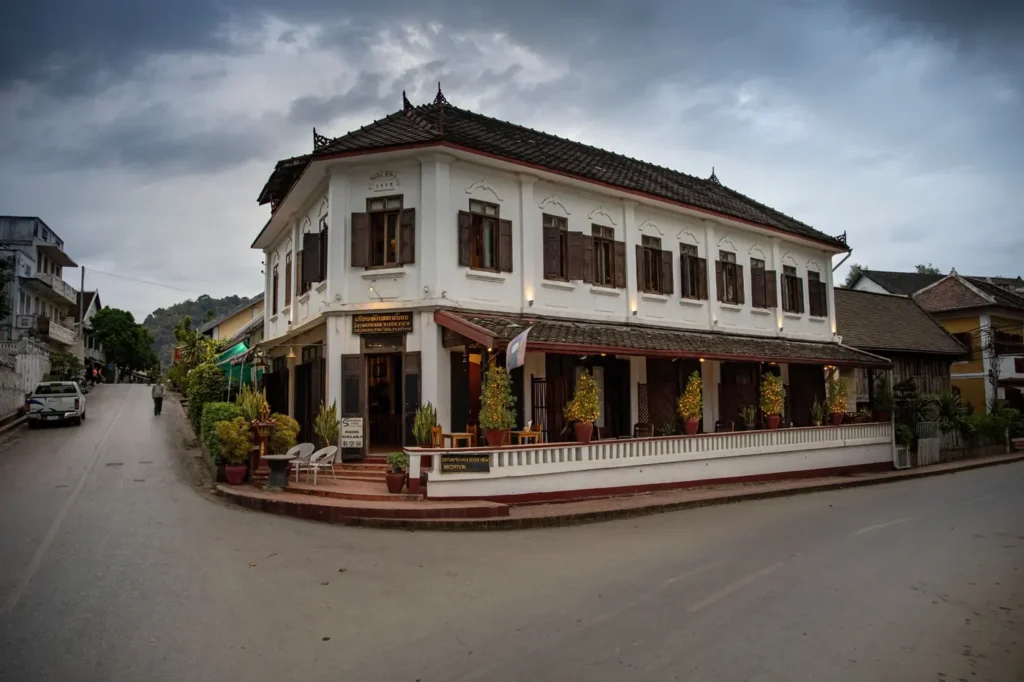 Alt text: Colonial-style boutique hotel on a quiet street with wooden shutters, potted plants, and a cloudy sky. 