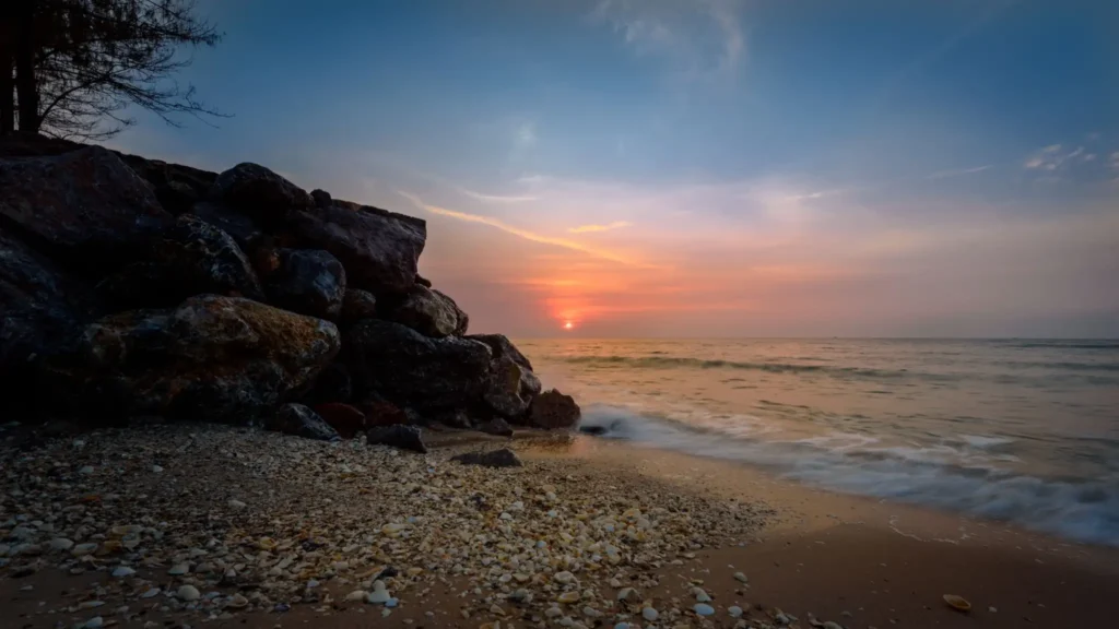 Rocky beach at sunset with gentle waves, seashells on the sand, and a warm orange sky over the ocean.