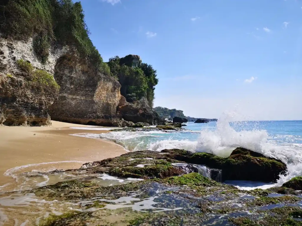 Rocky coastal beach cove with crashing waves, moss-covered rocks, sandy shoreline, and limestone cliffs under a clear blue sky
