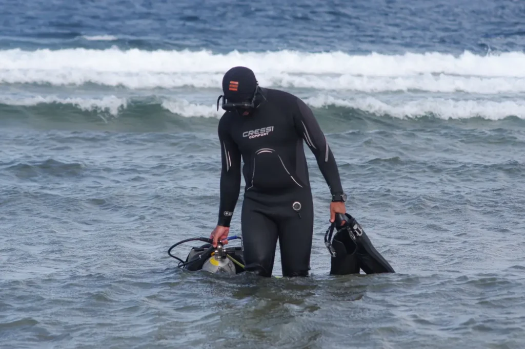 Scuba diver in shallow ocean water carrying diving gear near the shore.
