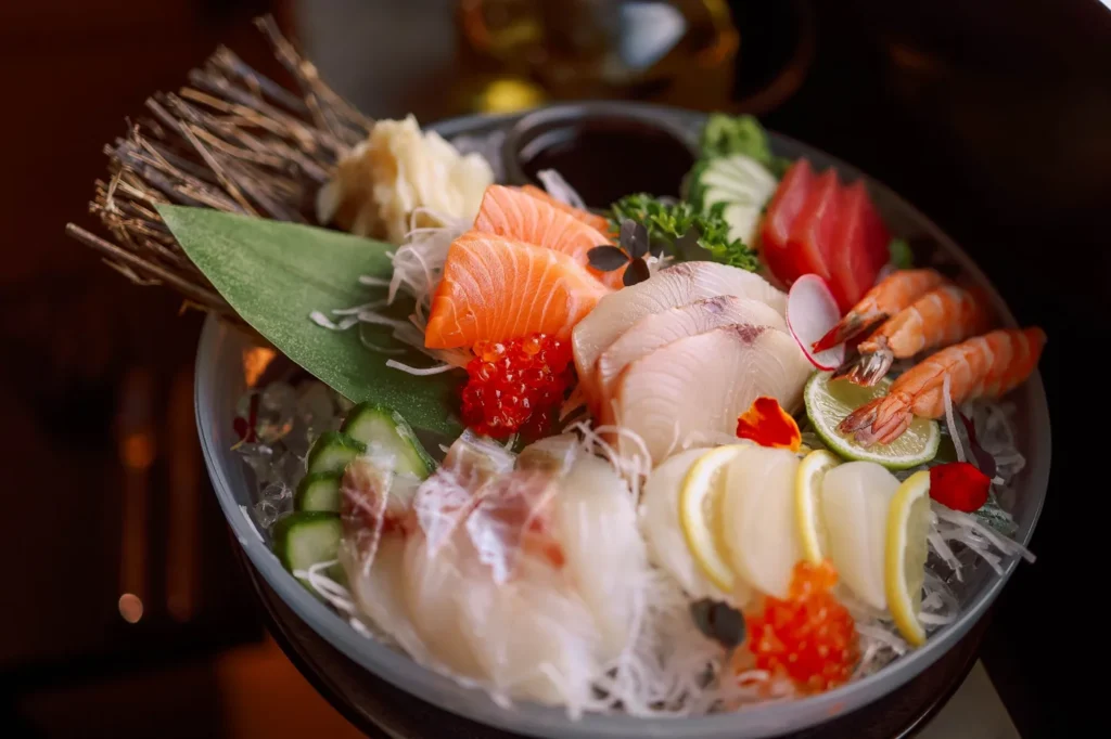 Fresh seafood sashimi bowl with salmon, white fish, shrimp, roe, lemon slices, cucumber, and garnish. 