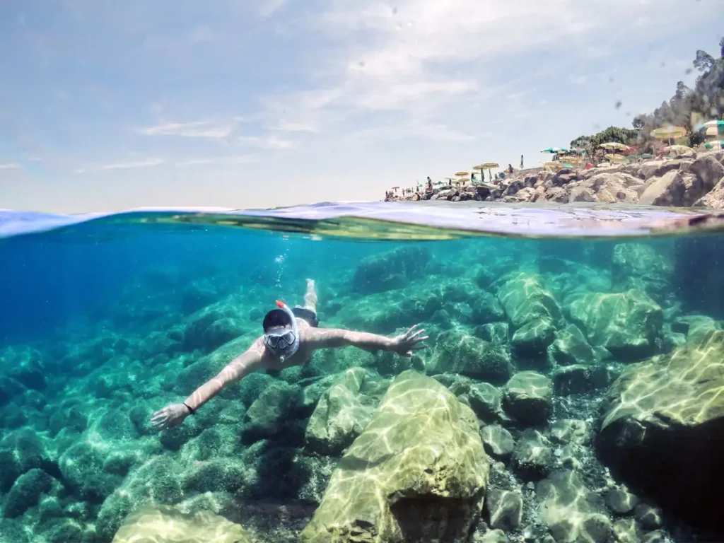 Snorkeler swimming above a rocky reef in crystal-clear turquoise water near the shoreline.