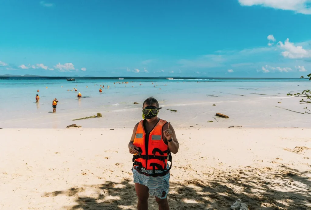 Person wearing a snorkel mask and life jacket on a white sandy beach with clear shallow water and swimmers in the background.