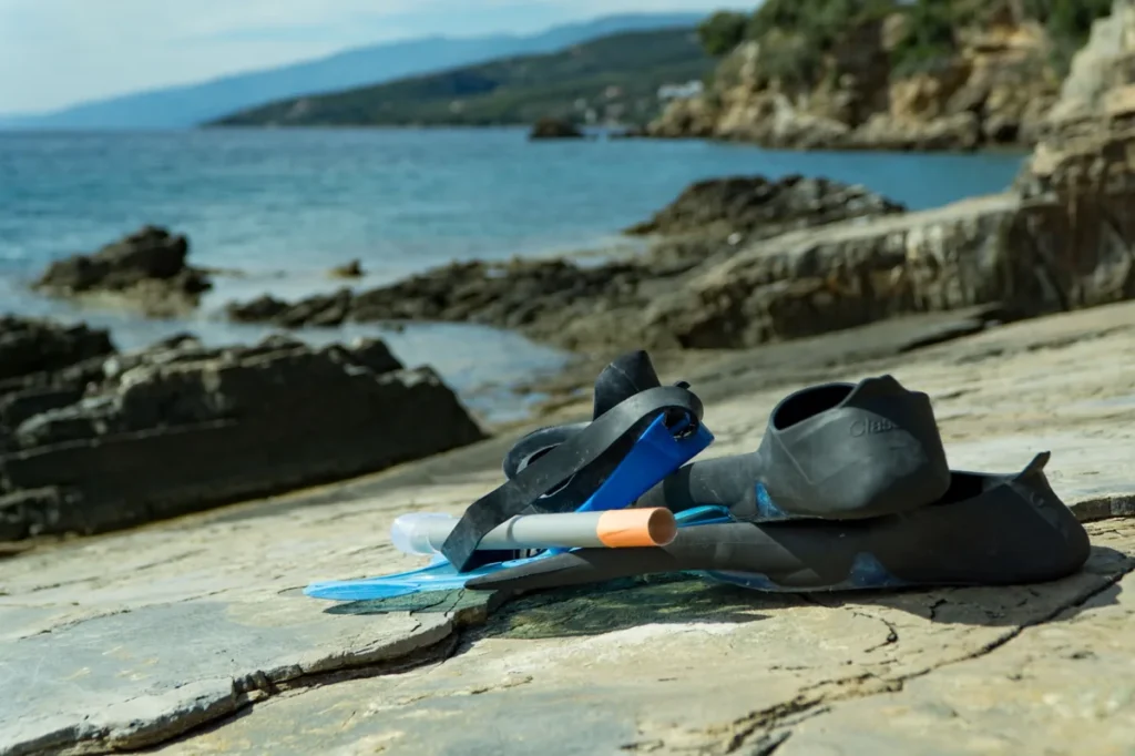 Snorkeling fins, mask, and snorkel placed on a rocky shoreline beside calm blue sea.