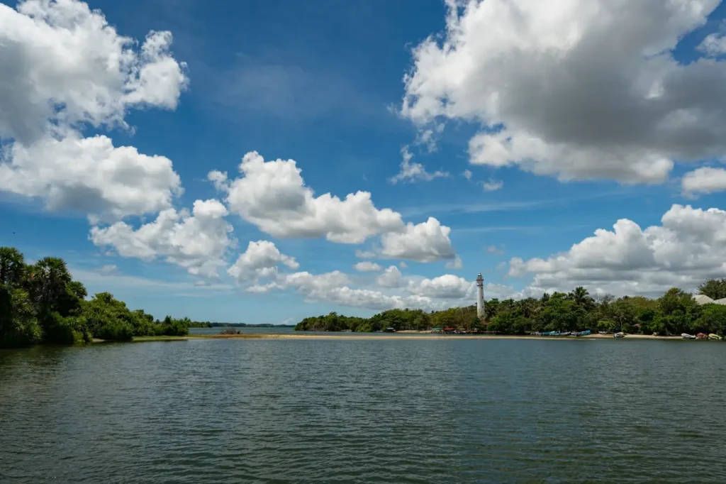 Calm Sri Lankan lagoon with a distant lighthouse, tropical trees, boats, blue sky, and large white clouds.