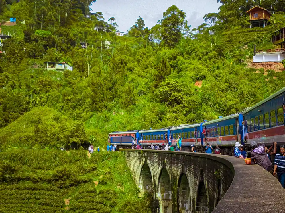 Blue train crossing a stone bridge through lush green hills in Sri Lanka with travelers enjoying the scenic railway view.
